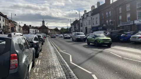 LDRS Yarm High Street with shops and residential buildings as well as numerous cars parked on either side of the road. There is traffic travelling in both directions.