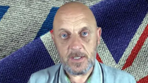 A bald and bearded man with blue eyes sitting in front of a Union flag