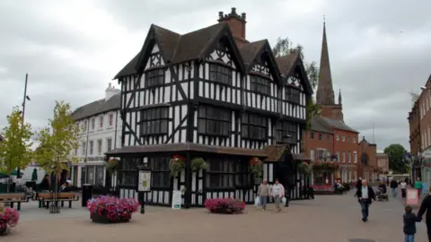 BBC A black-and-white Jacobean timber-framed house, Old House Museum, in Hereford. It is surrounded by planters full of colourful flowers and there is a church spire in the background.