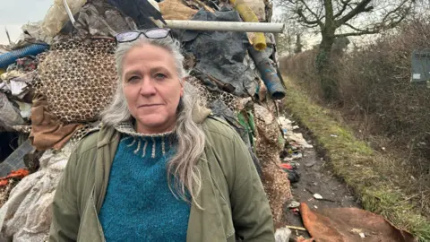 Caroline Farnell wears a turquoise jumper and a green coat over the top. She has glasses perched on top of her head and her hair is long and grey. She is standing in front of a huge pile of rubbish which towers above her.