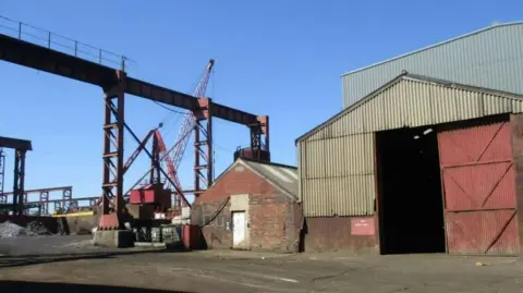 A metal recycling plant comprising of three buildigs made form stone and corrugated metal and a large piece of machinery painted red. 