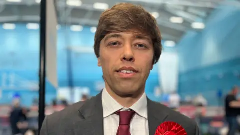 Emma Baugh/BBC Head and shoulders image of Cameron Holloway, standing in a sports hall. He has brown hair and is wearing a grey suit, red tie and white shirt.