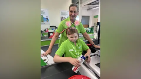 Asda Two people in green Asda uniforms stand at a supermarket checkout. One person is operating the barcode scanner and holding a tube of Colgate toothpaste over the scanning area, while wearing a pink headband decorated with colourful pom-pom-style antennae. The second person stands behind the checkout with both hands resting on the counter.
