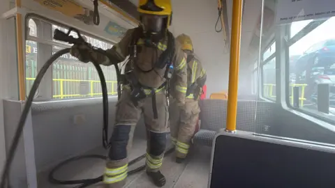 The inside of a Metro carriage is filled with smoke. Two firefighters, wearing bright yellow helmets with their visors down, are working inside.