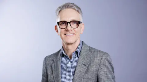 Falk Foundation eV A man with short grey hair is wearing black-rimmed glasses and smiling. He is wearing a shirt with small blue and white checks and a grey jacket