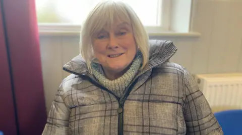 A woman in a checked coat wearing a roll-necked jumper with grey hair looks straight into the camera