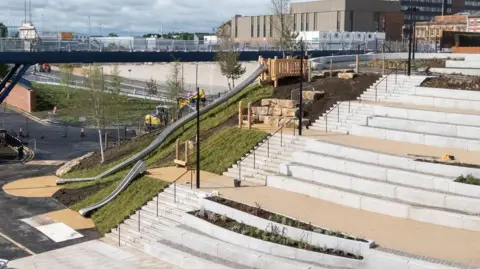 Stockton Council A long stainless steel metal slide runs down a grassy area. There are granite steps on the right and a footbridge in the background heads off over the river out of shot.