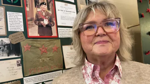 Linda wears glasses. She's standing with war memorabilia behind her. 