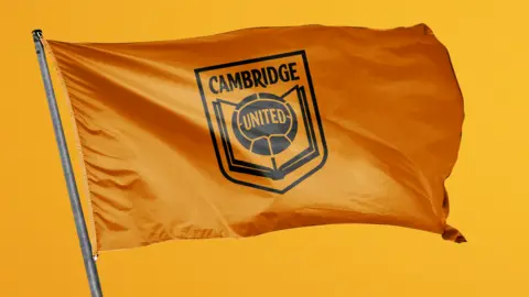 Cambridge United CUFC printed in black on an orange flag with a book and ball in the centre