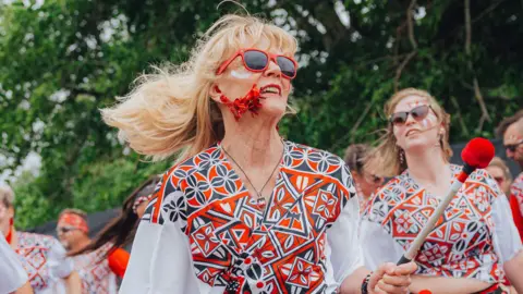 Jessie Myers A woman with sunglasses and patterned outfit in red, white and blue holds a stick for a drum.