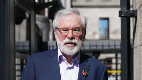 PA Gerry Adams with short grey hair and facial hair. He is wearing a navy suit with a light purple shirt. He has round, orange glasses on and there are badges on his suit jacket. Behind him is black gates.