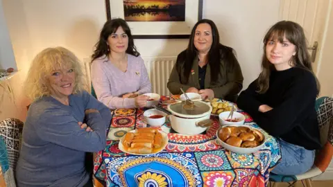 Four women sat inside a house around a table with plates of food and a brightly coloured, patterned table cloth. One woman has blonde hair and a blue top, one has black hair and a pink top, one has black hair and a dark green top and one has black hair and a black top