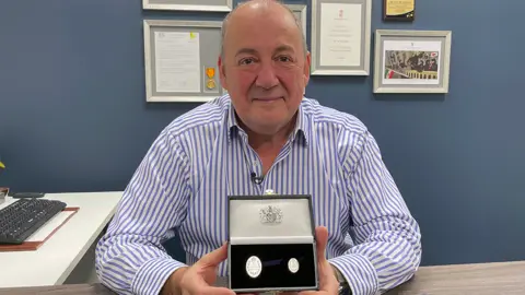 BBC Phil McDermott, in a blue striped shirt holding an Elizabeth Emblem medal in a case. He is sitting in an office with blue walls and a computer to his right