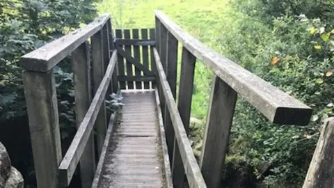 Bradford Council A wooden footbridge, leading to a grassy area. It is in a state of disrepair, with part of the bottom missing and weeds growing along the sides.