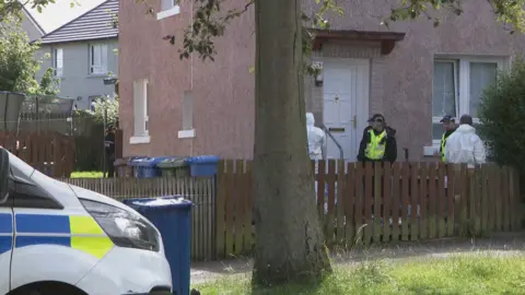 Police and forensic investigators in white boiler suits outside a house. A police car is parked outside on the road.