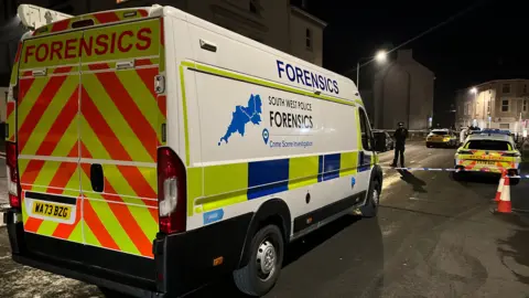 A forensics van on the left with police tape cordoning off a road. There are several police cars behind the tape and a police officer on the left. The photo was taken at night so the road is lit up by street lights. 