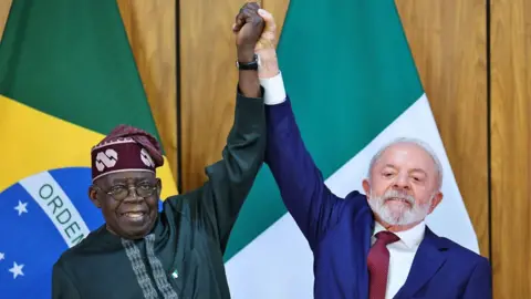 Adriano Machado/Reuters Brazil's President Luiz Inácio Lula da Silva, a blue suit, white shirt and maroon tie, and Nigeria's President Bola Tinubu, wearing a green outfit and maroon hat, raise their hands during a signing ceremony at the Planalto Palace in Brasília, Brazil - Monday 25 August 2025. 