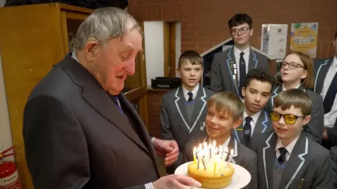 BBC Dr Neville Brown berdiri dalam setelan jas di sebelah kiri gambar memegang kue dengan lilin, dengan sekelompok selusin siswa Maple Hayes berdiri di sebelahnya.