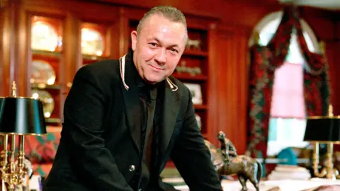 David Sullivan wears a black suit while sitting on a desk in his home in 2003. 