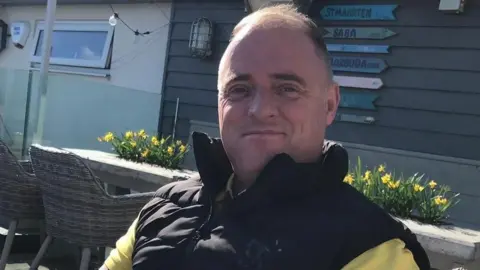 Supplied Ray McDonald is sitting on an outdoors chair with a house or cabin and a row of daffodils behind him. He is about 50, clean-shaven with a receding hairline and is wearing a black sleeveless jacket over a yellow top.
