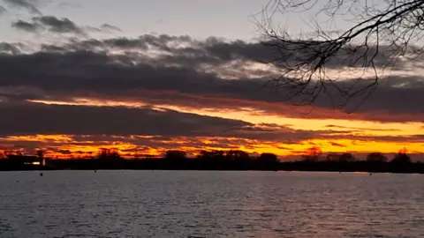 Nigel Wise A deep orange sky with dark clouds around it seen over a pinkish lake reflecting the sunset. 