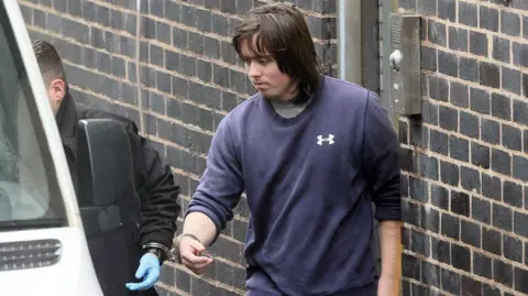 Daily Post A man with long brown hair, wearing a blue sweatshirt. he is attached to a guard wearing a black jacket by handcuffs and is being led into a white van.