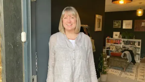 A woman is standing at her open front door. She is smiling. Behind her we can see the lounge.