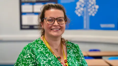 Martin Giles/BBC A teacher wearing a green and white floral dress is smiling at the camera. She has brown hair and is wearing turquoise glasses. She has several necklaces on and is wearing a yellow lanyard.