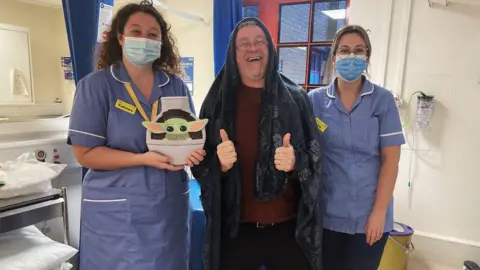 Inspiring Heritage Mr Hale in hospital with two nurses beside him, while he was recovering from bowel cancer. He is smiling and giving two thumbs up for the camera. He is dressed as a Jedi and one of the nurses is holding a baby Yoda hot water bottle. 