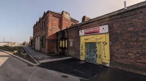 Thomas Ollivier The exterior of the Strand Railway Station and club in Barrow. The building has old bricks and is boarded up. It has graffiti on some of its exterior and a red sign reading 'RAILYWAYMENS CLUB' above what was once a door. The other parts of it has been boarded up.