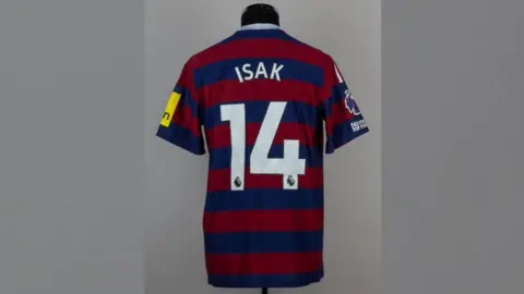 Graham Budd Dark red and dark blue striped Newcastle United shirt which says Isak and the number 14. It is on a mannequin.