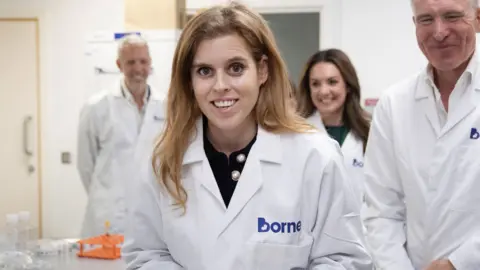 Princess Beatrice during a visit to Borne's laboratories. She is wearing a lab coat and smiling at the camera