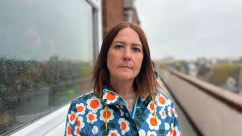 Katie Courtney-Jones standing outside flats in Guildbourne Court on a walkway on the top floor next to a window. She has brown hair and a flowery rain coat
