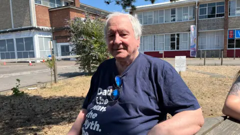 Clare Worden/BBC A man with white hair sits on a wooden bench in front of a building. He has sunglasses tucked into his blue T-shirt. 