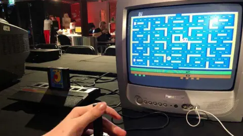 Image shows hand at the controls of an Atari game console playing Pac-Man on a Panasonic television