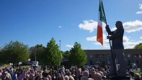 PA Media A crowd of between 50 and 100 people stand around a statue of hunger striker Bobby Sands holding a tricolour in a housing estate in Belfast