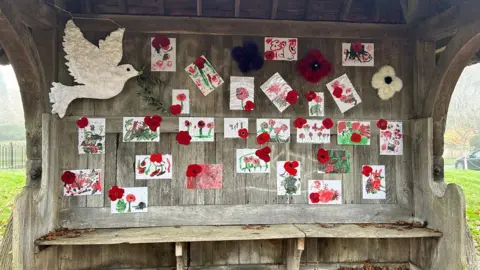 Holt Residents A bench with wooden shelter and back with a large dove of peace in one corner and childrens pictures of poppies, knitted ones scattered in between