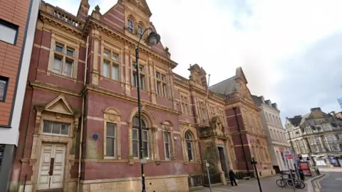 General view of a late 19th-Century red-brick building in Wolverhampton city centre. It has arched windows and other features in paler stone, and sits in a city streetscape between buildings of different designs and eras. 