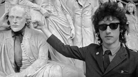 Getty Images Clarke standing next to a statue of William Shakespeare with a finger in one of the statue's ears, at the Prince Albert Memorial in Hyde Park, London, in 1978