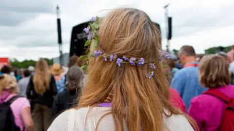 A woman is wearing a ring of flowers around her head. She is standing in a crowd of people at a festival, looking at a large stage with two tall speakers on either side.