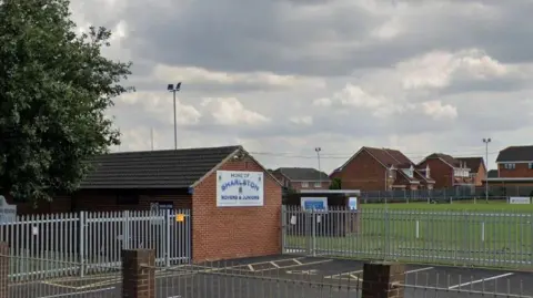 Google The outside of a rugby clubhouse surrounded by metal gates and a sports pitch in the background.