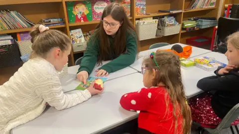 A girl with long dark hair and a green jumper sits at a low table in-front of three much younger pupils who are all looking down at a book.