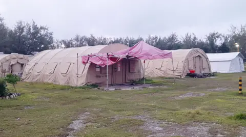 Deretan tenda besar berwarna khaki tempat para migran ditampung secara berkelompok, dengan beberapa tenda berwarna putih yang digunakan sebagai ruang komunal darurat.