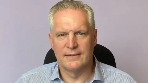 Dr Stout has short grey hair and is wearing a blue and white striped shirt.