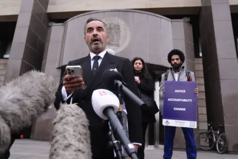 PA Media Lawyer Aamer Anwar stands in front of Glasgow Sheriff Court, speaking into a number of microphones. He is wearing a black suit, black tie and white shirt. Standing behind him are two campaigners, a man and a woman. The man is holding a sign which says "justice, accountability, change".