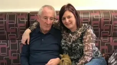Michael Griffiths, with short white hair and glasses, wearing a blue sweater, is sitting alongside Brenda Brereton, who has long brown hair and is wearing a brown and white patterned top. She has her right arm around her father's shoulders. They are sitting on a brown sofa with a red and white pattern.