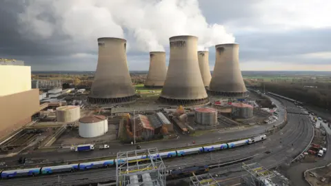 A power station with five large chimneys. Low rise buildings around the station and a train running around the outside.