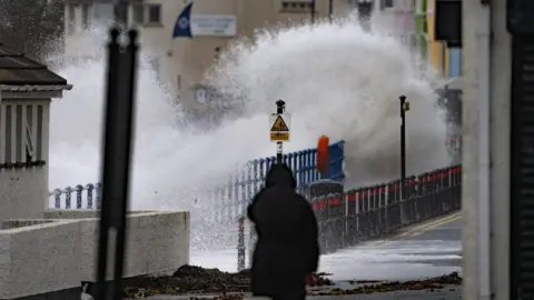 A person wearing a black hooded coat is walking towards a street, where a wave is crashing over a fence. Terraced houses painted in different colours are across the street.