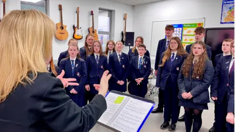 Martin Giles/BBC A woman leads a choir of teenage boys and girls dressed in navy school uniform in a music room, with guitars hanging on the walls