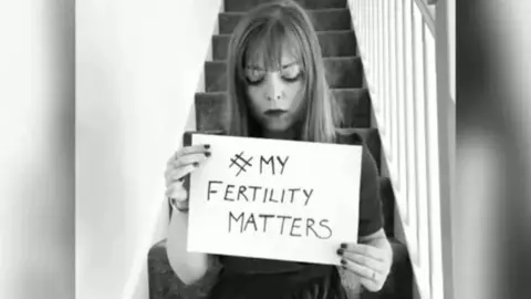 Sarah Curtiss A black and white photo of Sarah Curtiss sitting on stairs in a home and holding a hand-written sign reading "#My Fertility Matters".